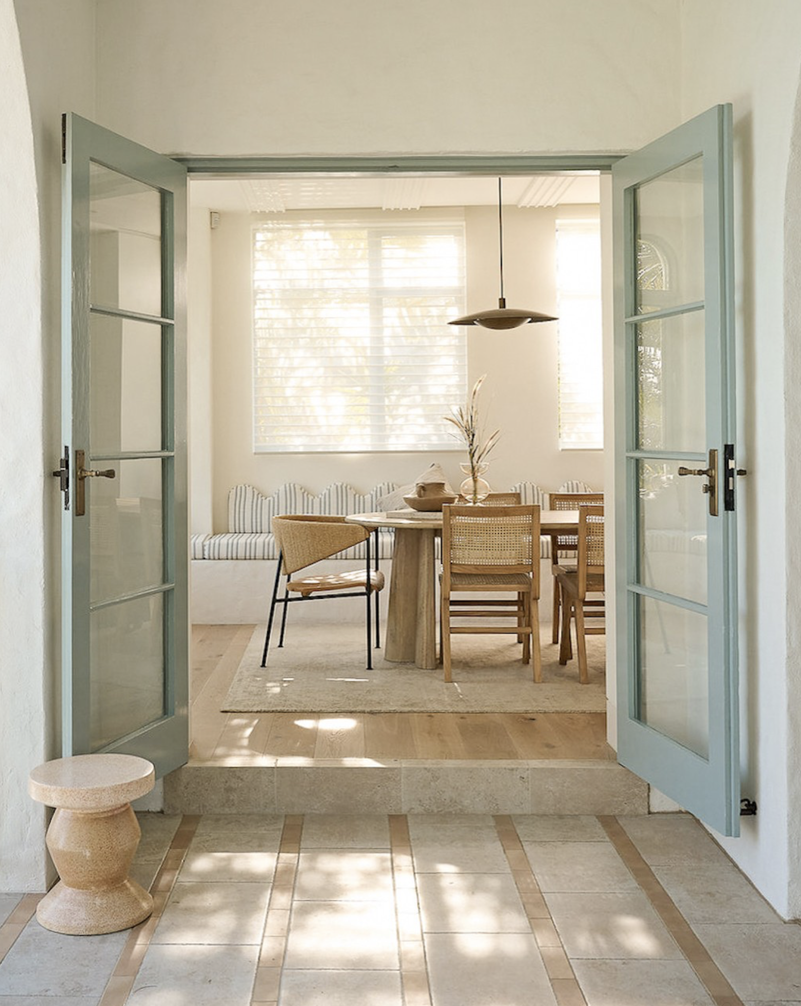 Dining room interior featuring a round wooden table, woven chairs, and natural light through open doors and window blinds.