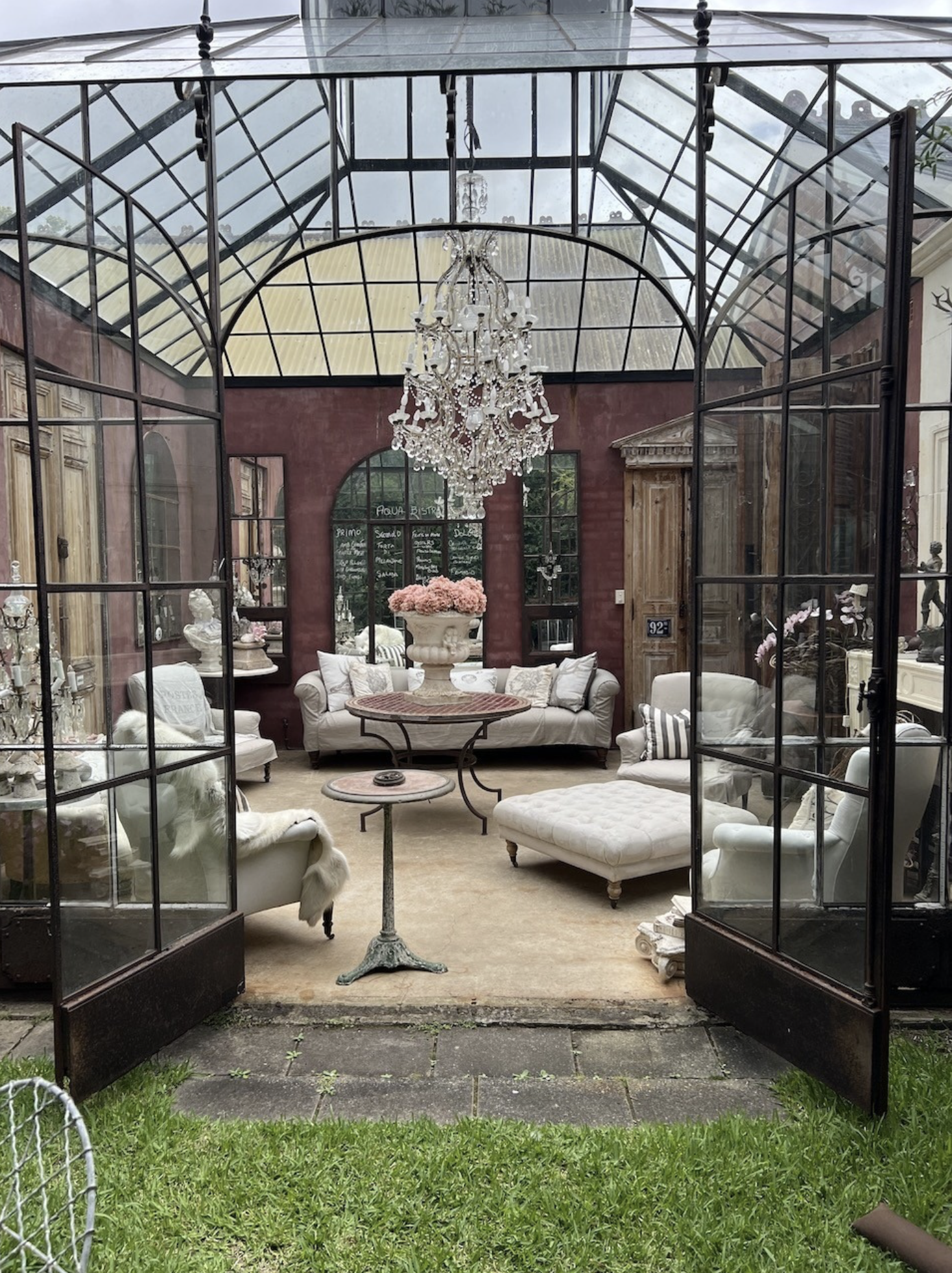 Ornate sunroom with black metal framing, glass ceiling, crystal chandelier, neutral furniture, and garden view through open doors.