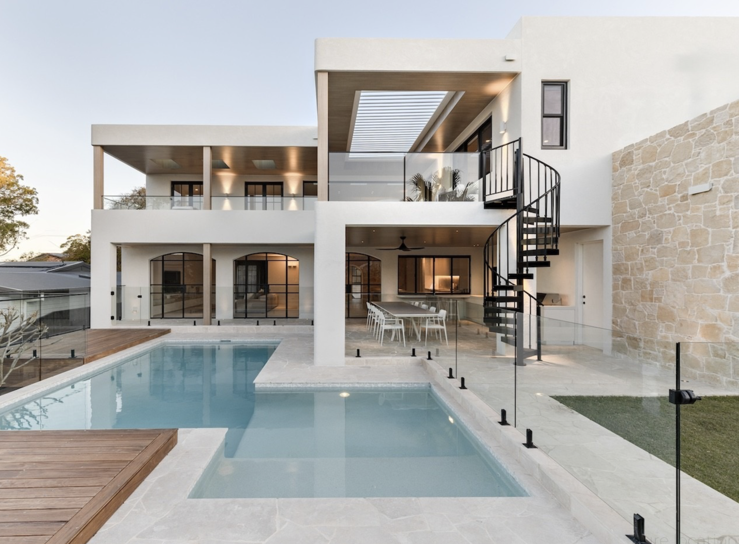 Exterior of a modern two-story house with a swimming pool, patio dining, and spiral staircase, surrounded by glass fencing and stonework.