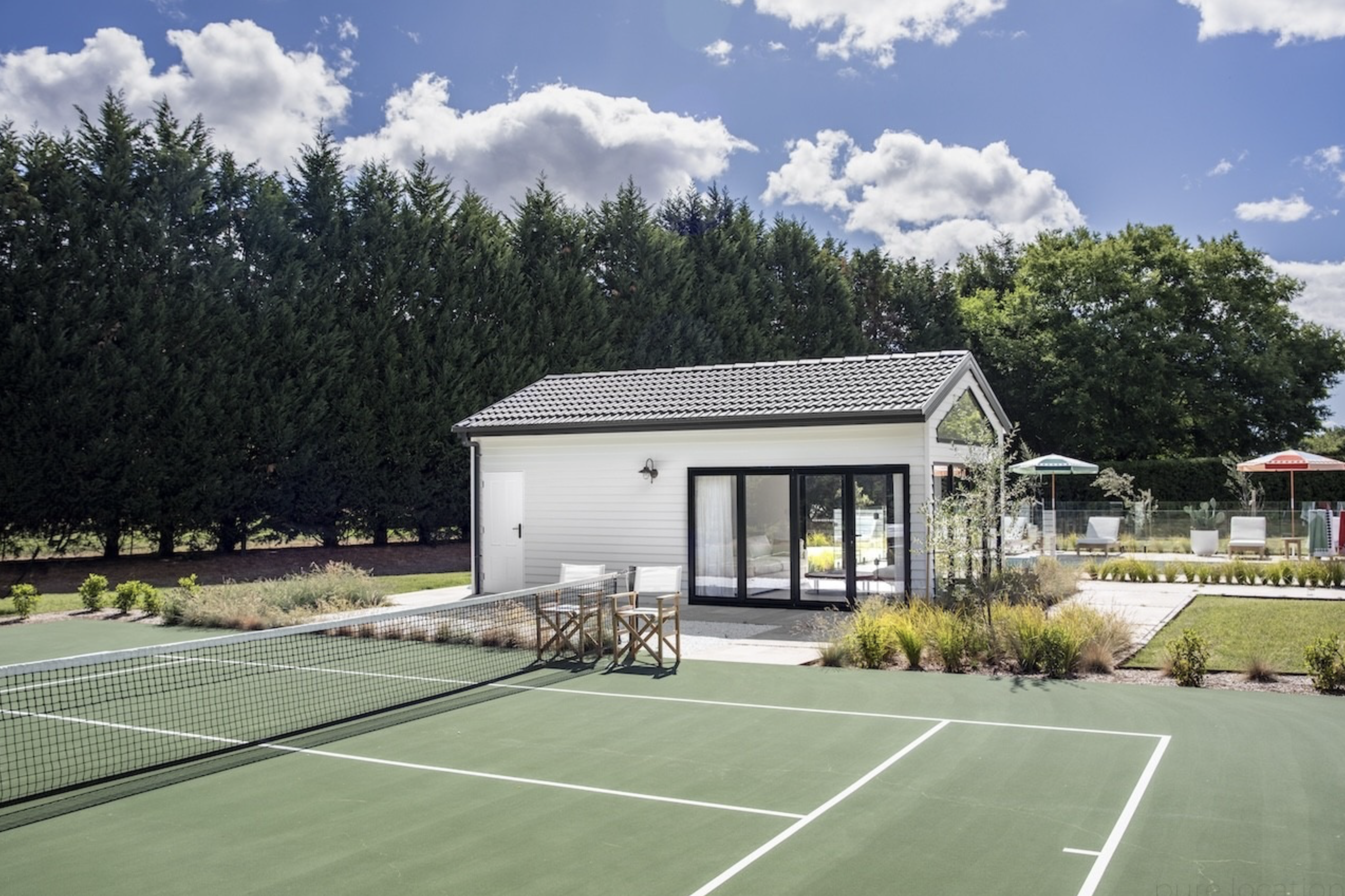 Exterior view of a tennis court with a green surface, white lines, a net, a white service building, and green trees and blue sky.