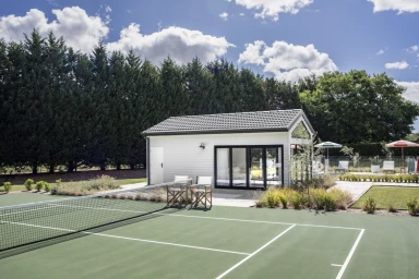 Tennis court with white net in foreground and a white modern garden studio cabin set against a tall green hedge and bright sky.