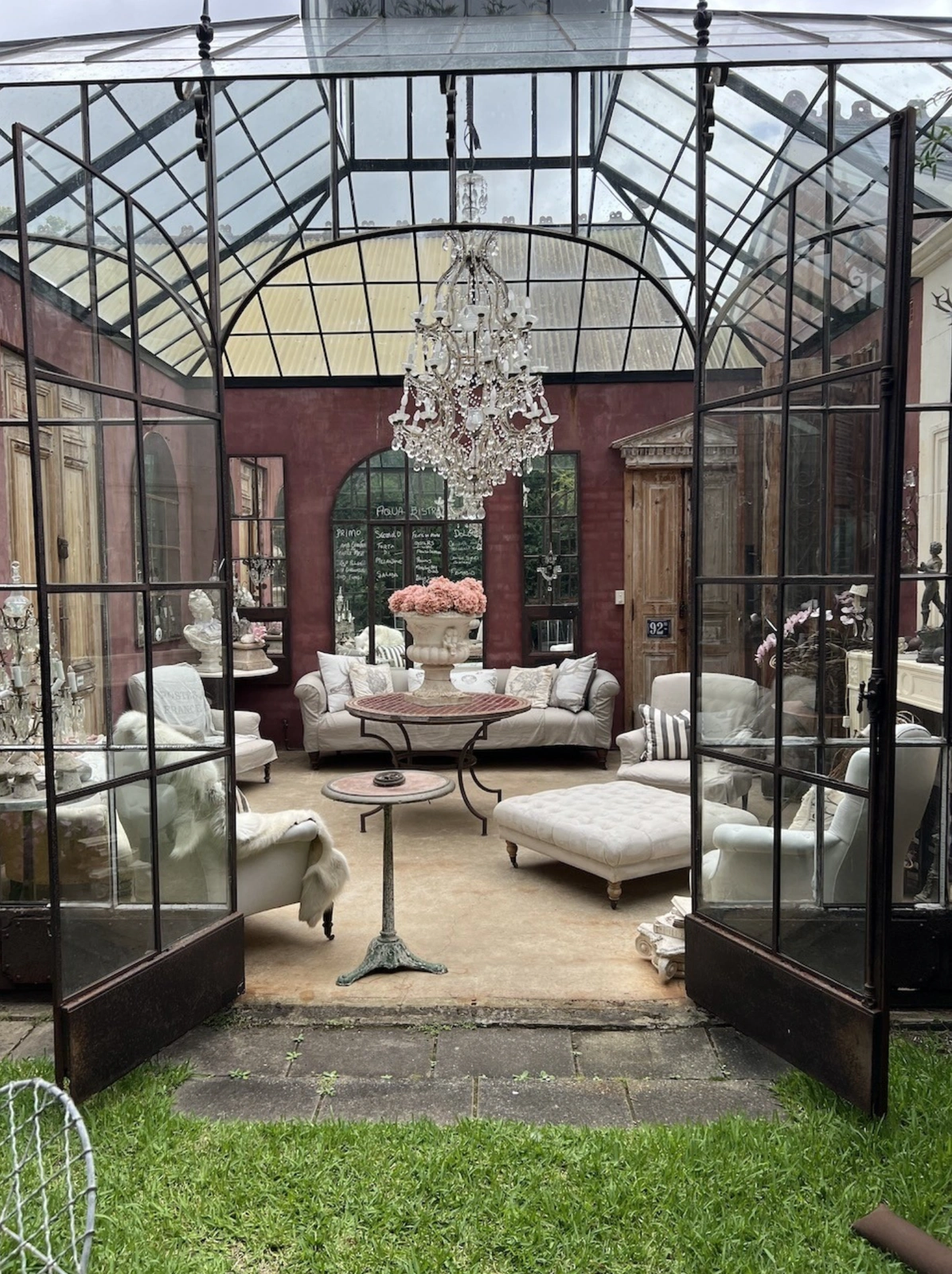 Glass conservatory sunroom featuring a crystal chandelier, neutral sofas, tufted ottoman, and arched mirrors by Pure Locations Sydney