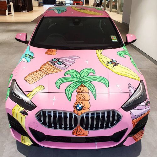 Eye-level shot of a pink BMW car featuring a cartoon-style paint job with images of bananas, ice cream cones, and palm trees on the hood.
