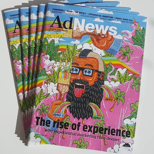 Stack of AdNews magazines on a white surface with a cartoon-style cover featuring a man with a beard and pink palm tree shirt.