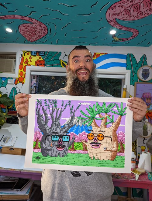 Bearded man holds an art print of two cartoon trees wearing glasses against a vibrant, colorful studio background with painted walls.