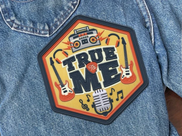 A close-up of a denim jacket featuring a vibrant music-themed patch. The patch showcases retro audio equipment and the words 'True To Me'.