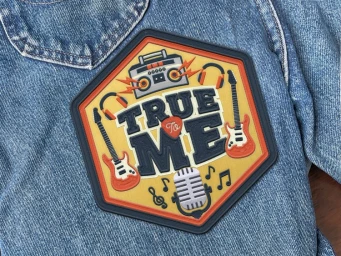 Hexagonal embroidered patch with the words True To Me surrounded by guitars and a boombox on blue denim fabric.