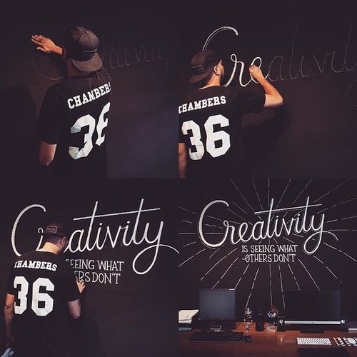 Man in black shirt writing 'creativity' on dark wall, wearing cap, seen from the back, calligraphy art style with desk and computers