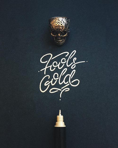 Still life of a skull ring, a calligraphy pen, and the script "Fools Gold" against a dark background.