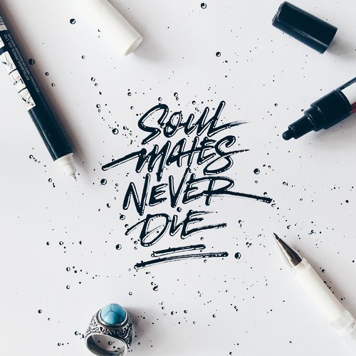 Calligraphy art featuring 'Soul Mates Never Die' with artistic ink splatter, pens, and a ring on a white background, top-down shot.