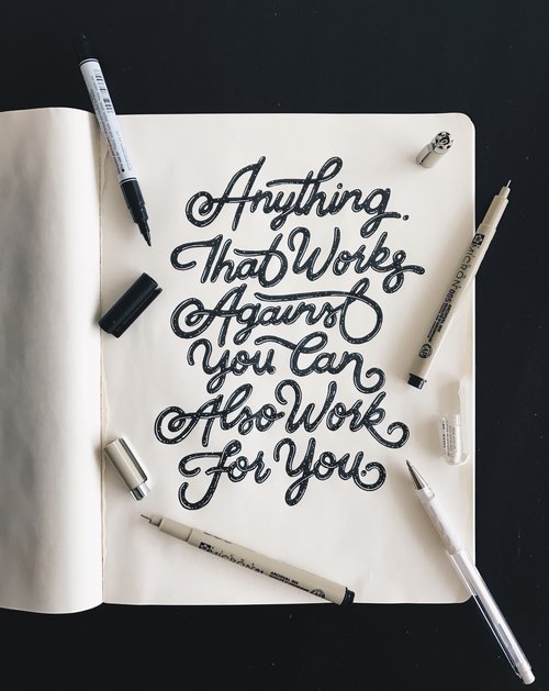 Open sketchbook with handwritten lettering, featuring various black marker pens; perfect for typography and creative design projects.