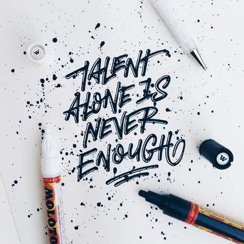 Close-up of black handwritten calligraphy, talent alone is never enough, with art markers and ink splashes on white paper background.