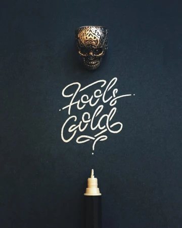 An ornate metal skull ring above white handwritten calligraphy text spelling Fools Gold and a white ink pen against a dark background