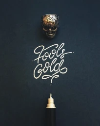 An ornate metal skull ring above white handwritten calligraphy text spelling Fools Gold and a white ink pen against a dark background