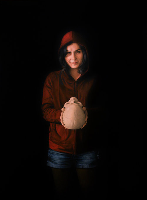 Portrait of a young woman in a red hoodie holding a clay skull against a dark background, lit with soft lighting.