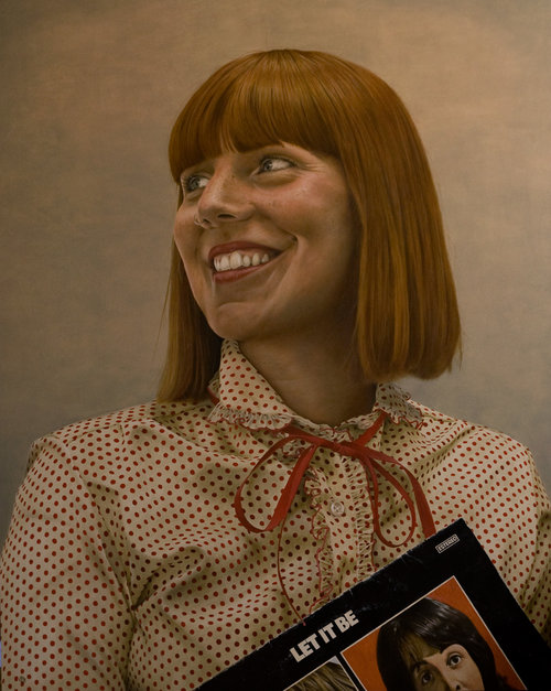 A woman with auburn hair smiles while holding a Beatles "Let It Be" album, posing indoors in front of a neutral background.