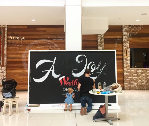 Adult male chalk artist creates mural of 'A Joy' on large blackboard with toddler daughter at Westfield Chatsworth mall in a commercial indoor setting.