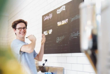 Smiling man wearing glasses and a grey t-shirt holding chalk in front of a dark menu board on a white tiled wall.