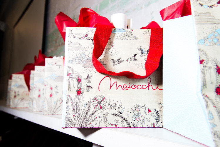 Close-up of several branded shopping bags with floral and bird designs, featuring red ribbon handles, set against a neutral wall.