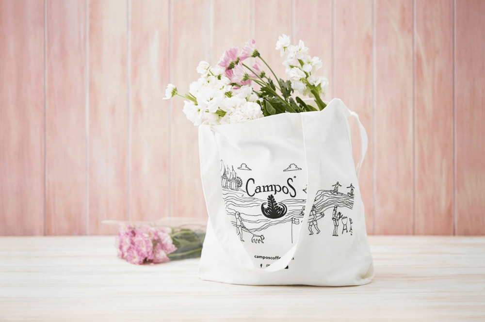 White tote bag filled with pink and white flowers, standing on a white table against a light pink wood-panelled background.
