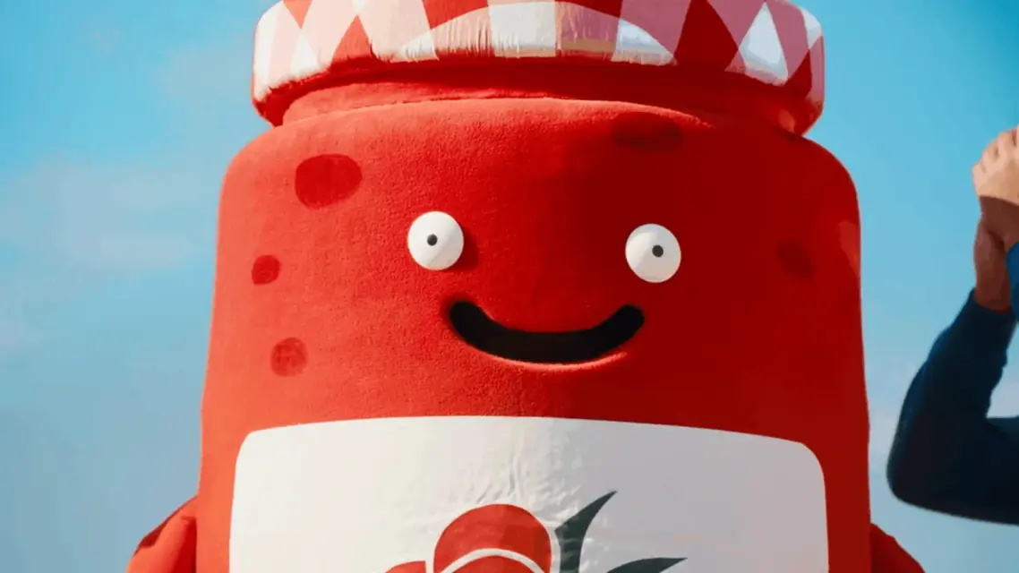 A vibrant red jar mascot character with a cheerful smile stands outdoors under a bright blue sky, adding a playful touch.