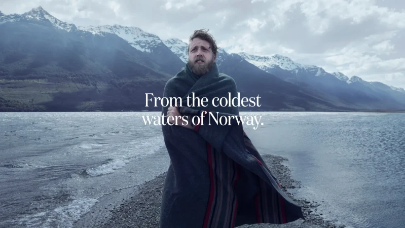Bearded man wrapped in a dark wool blanket standing on a stony shore with mountains and water in the background.