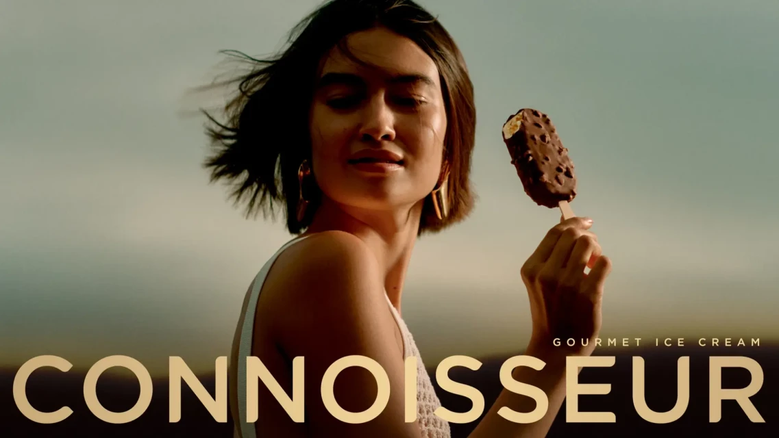 Woman in outdoor setting holds gourmet ice cream bar, promoting Connoisseur brand with visible logo and relaxed, sunlit ambiance.