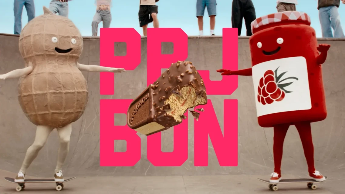 Humorous food costumes on skateboards against a skatepark backdrop, featuring a peanut, chocolate bar, and a jar of jam.