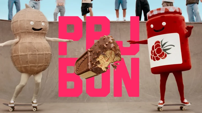 Humorous food costumes on skateboards against a skatepark backdrop, featuring a peanut, chocolate bar, and a jar of jam.