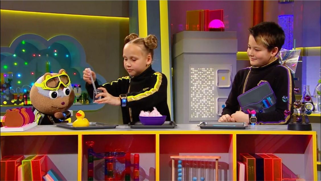 Two children and a puppet are in a television studio on a set with lab equipment and colorful decor, filming a science segment.