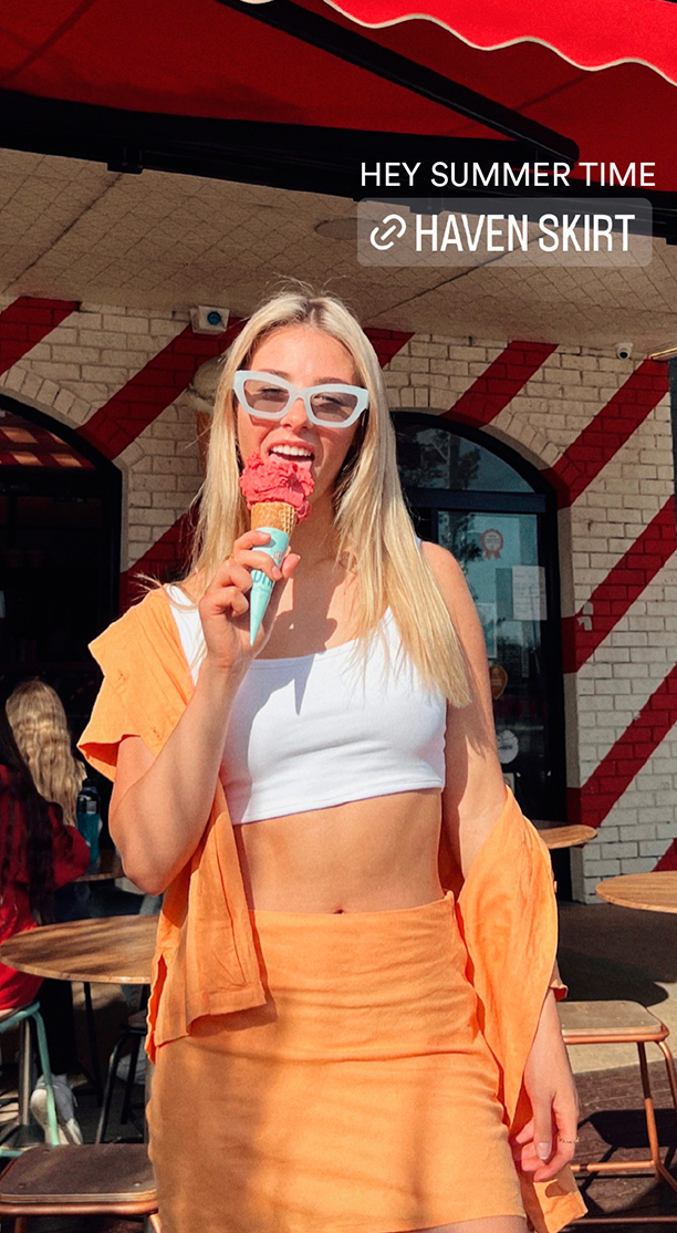 Blonde woman in orange skirt eating strawberry ice cream cone outdoors in front of a brick building with red and white awning.