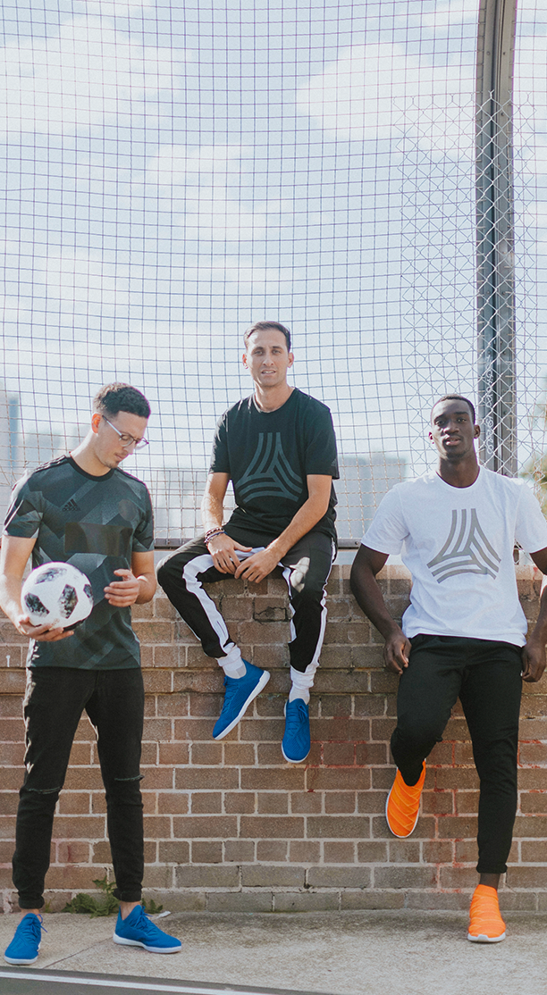 Three young adult men in athletic wear pose in front of a brick wall and chainlink fence, holding a soccer ball, outdoors in daylight.