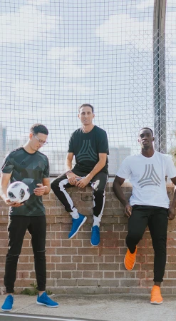 Three young adult men in athletic wear pose in front of a brick wall and chainlink fence, holding a soccer ball, outdoors in daylight.