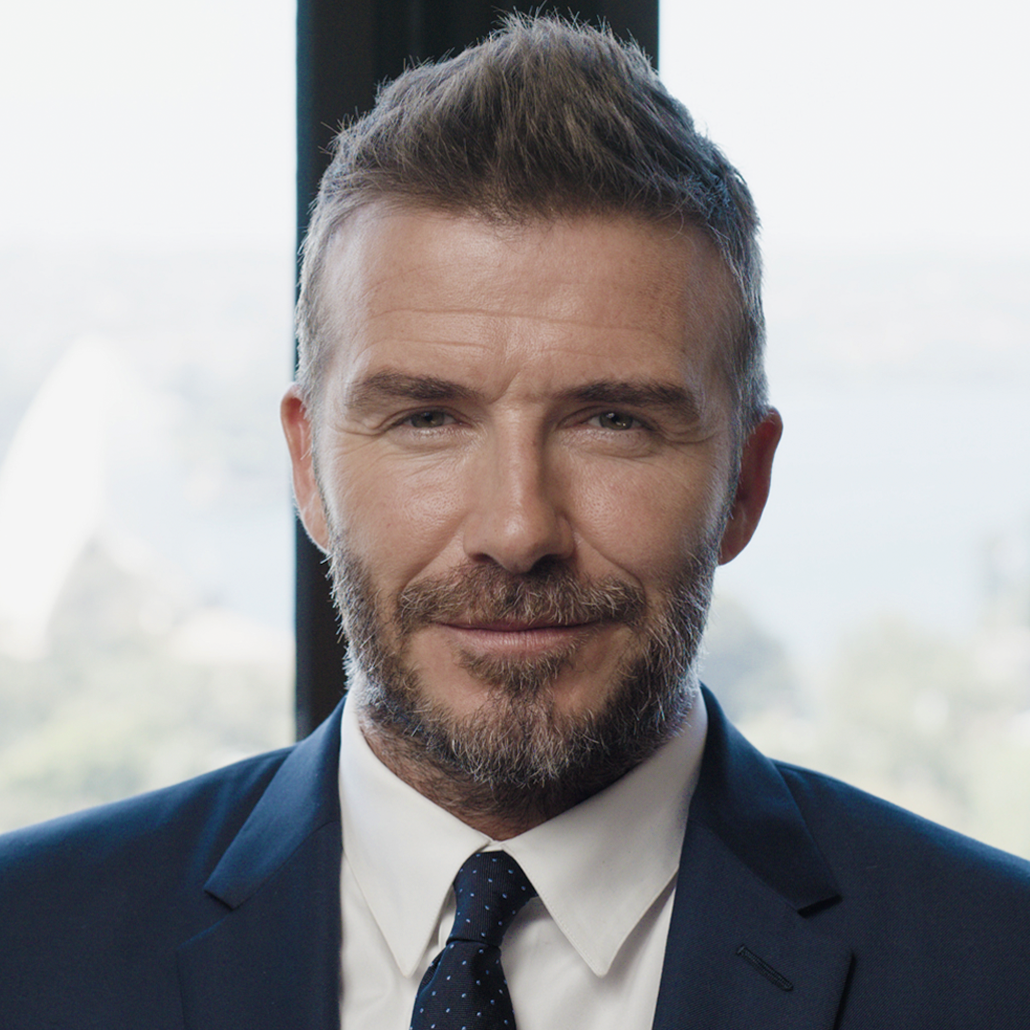 Close-up portrait of a smiling adult male in a dark suit and tie, indoors, with a blurred neutral-color background.