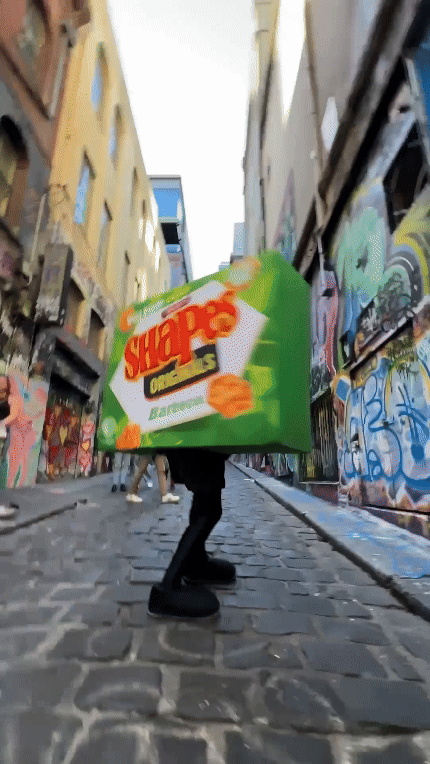 Street scene features a person in a snack food box costume walking down a narrow alley with graffiti walls, creating a surreal urban moment.