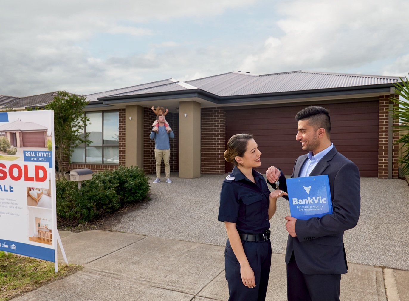 Real estate agents meet in front of house with sold sign as father and child stand in the doorway of a modern suburban home.