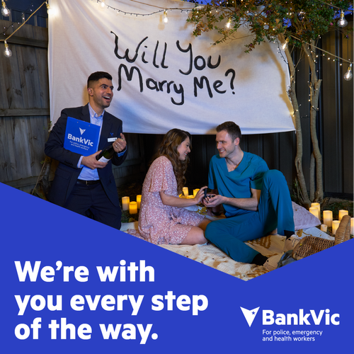 Outdoor proposal scene with a couple kneeling, a friend holding champagne, lit by candles and string lights, captured at eye-level.