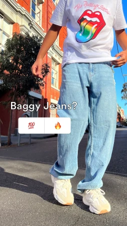 A young adult poses in baggy light blue denim jeans and white sneakers on an urban street with brick buildings. White t-shirt with Rolling Stones tongue.