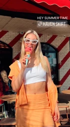 Blonde woman wearing white cat-eye sunglasses and orange skirt eating ice cream cone outdoors