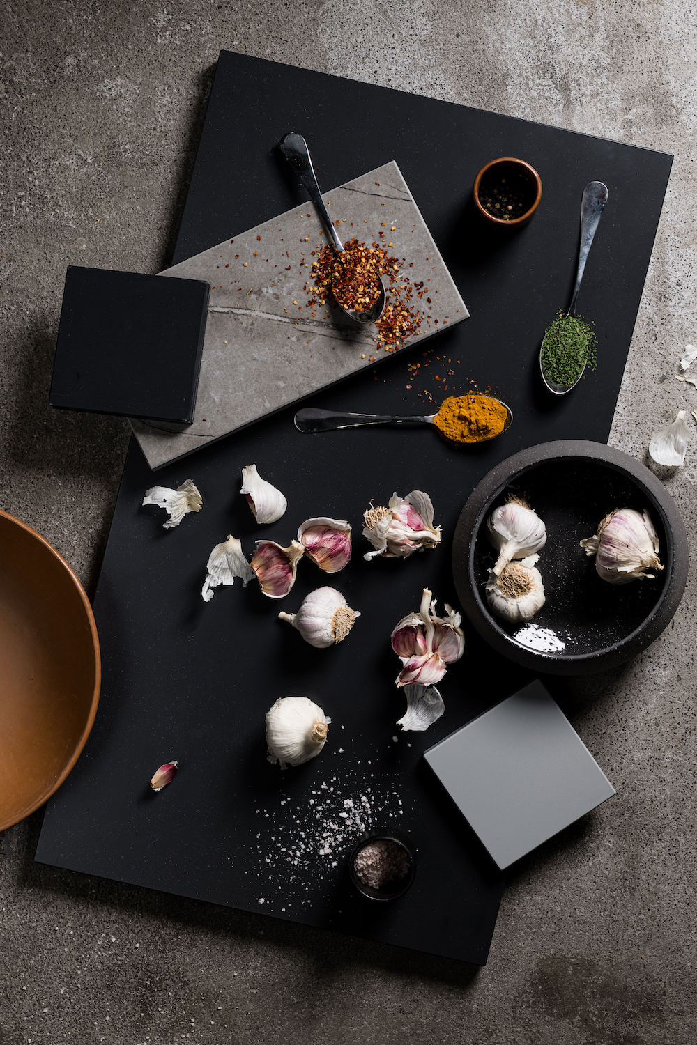 Overhead shot of culinary ingredients and spices arranged on square black and grey presentation boards, including garlic, salt, chili flakes.