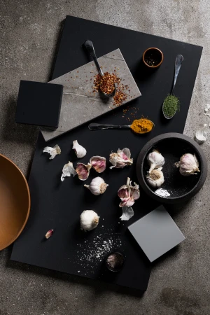 Overhead shot of culinary ingredients and spices arranged on square black and grey presentation boards, including garlic, salt, chili flakes.
