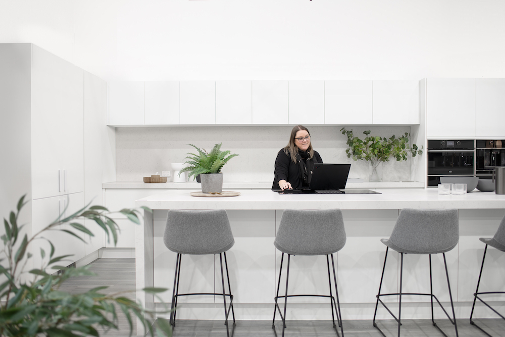 Woman with glasses works on a laptop in a modern, bright white kitchen with grey stools and potted plants on the island counter.
