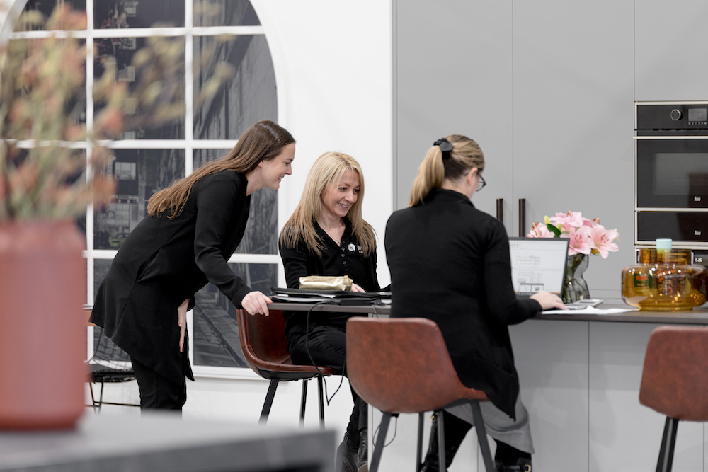 Three women are gathered at a table in a modern interior. One woman is seated at a laptop, another is seated while talking to her.