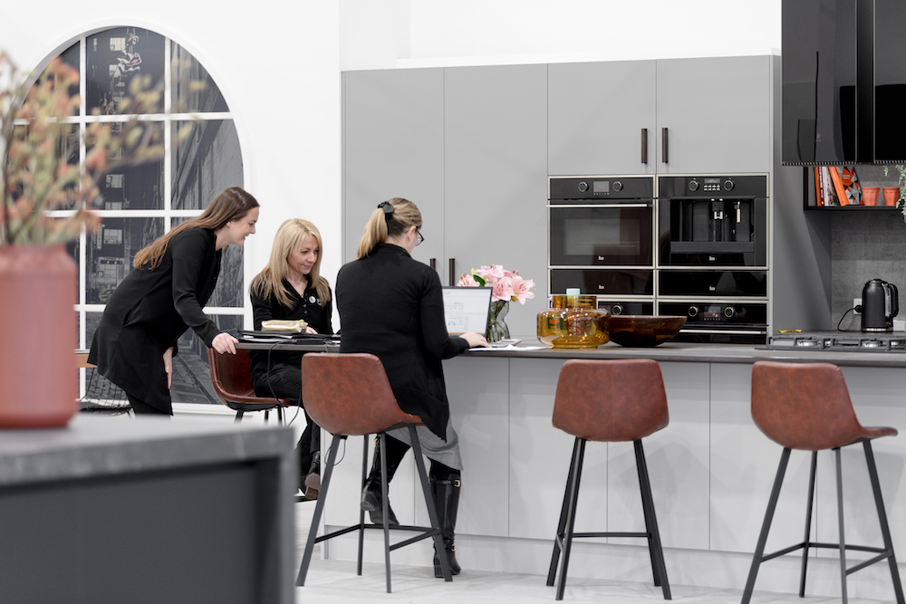 Three women in a modern kitchen with gray cabinets, dark counter, and brown leather bar stools; one woman using laptop at counter.
