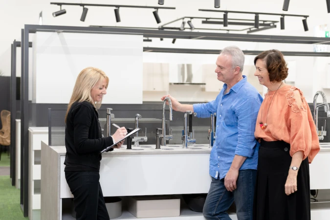 Couple shopping for kitchen fixtures with assistance from a saleswoman, standing in a showroom with modern faucet displays.