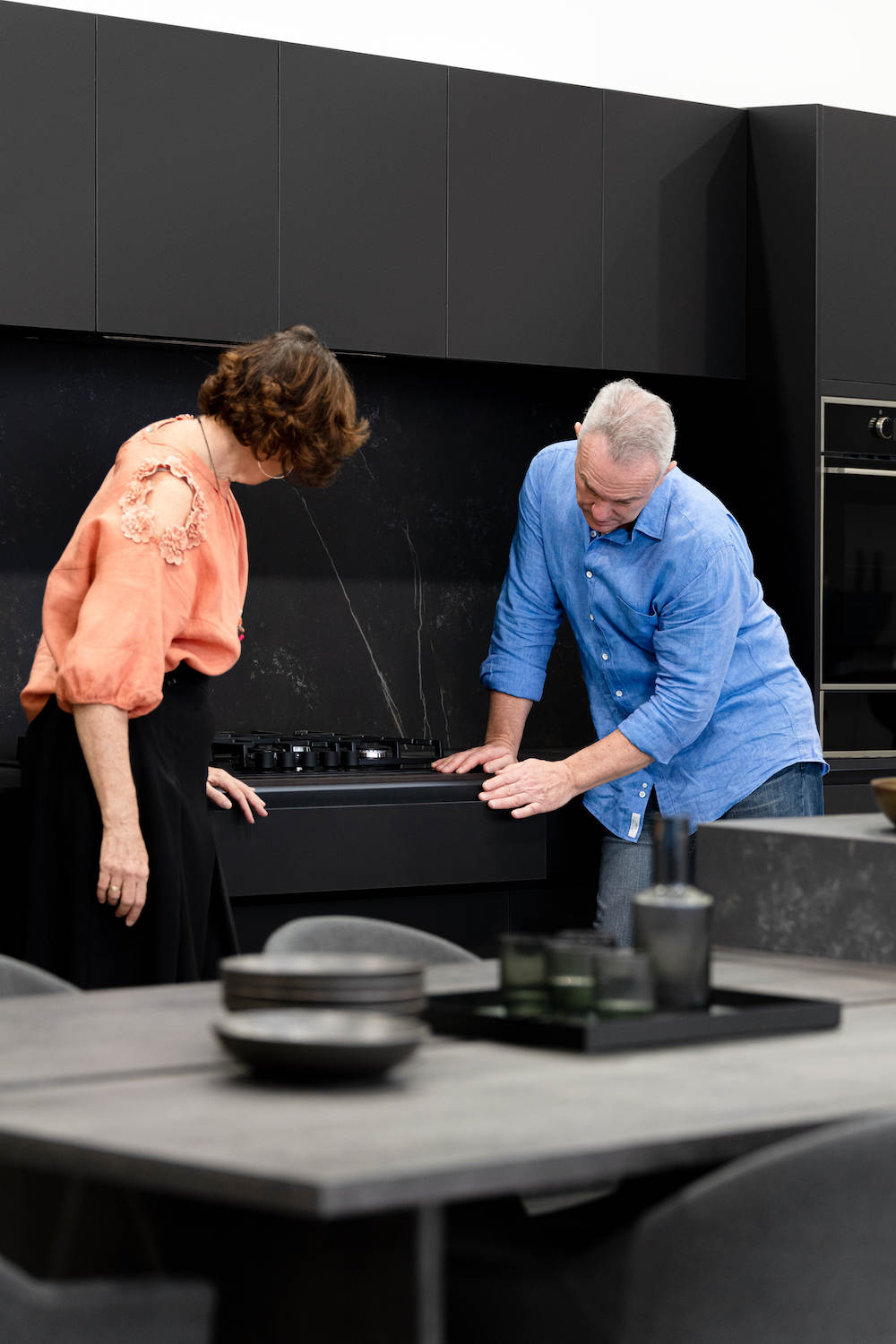 A modern black kitchen with a couple looking at the gas stovetop, dark wood table with dishes and glassware in the foreground.