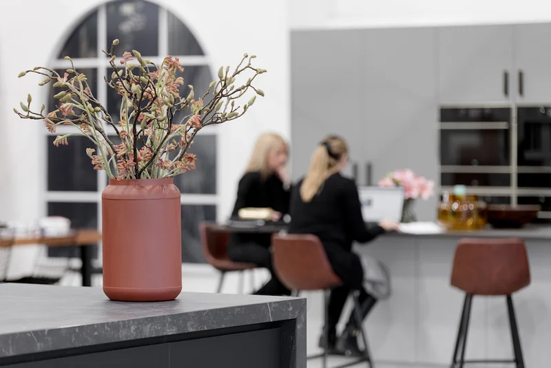 Terracotta vase with dried stems on a dark grey countertop with two people seated in a modern kitchen background.