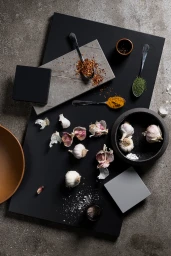 Overhead flat lay of garlic bulbs, scattered spices, and material samples arranged on a black board.