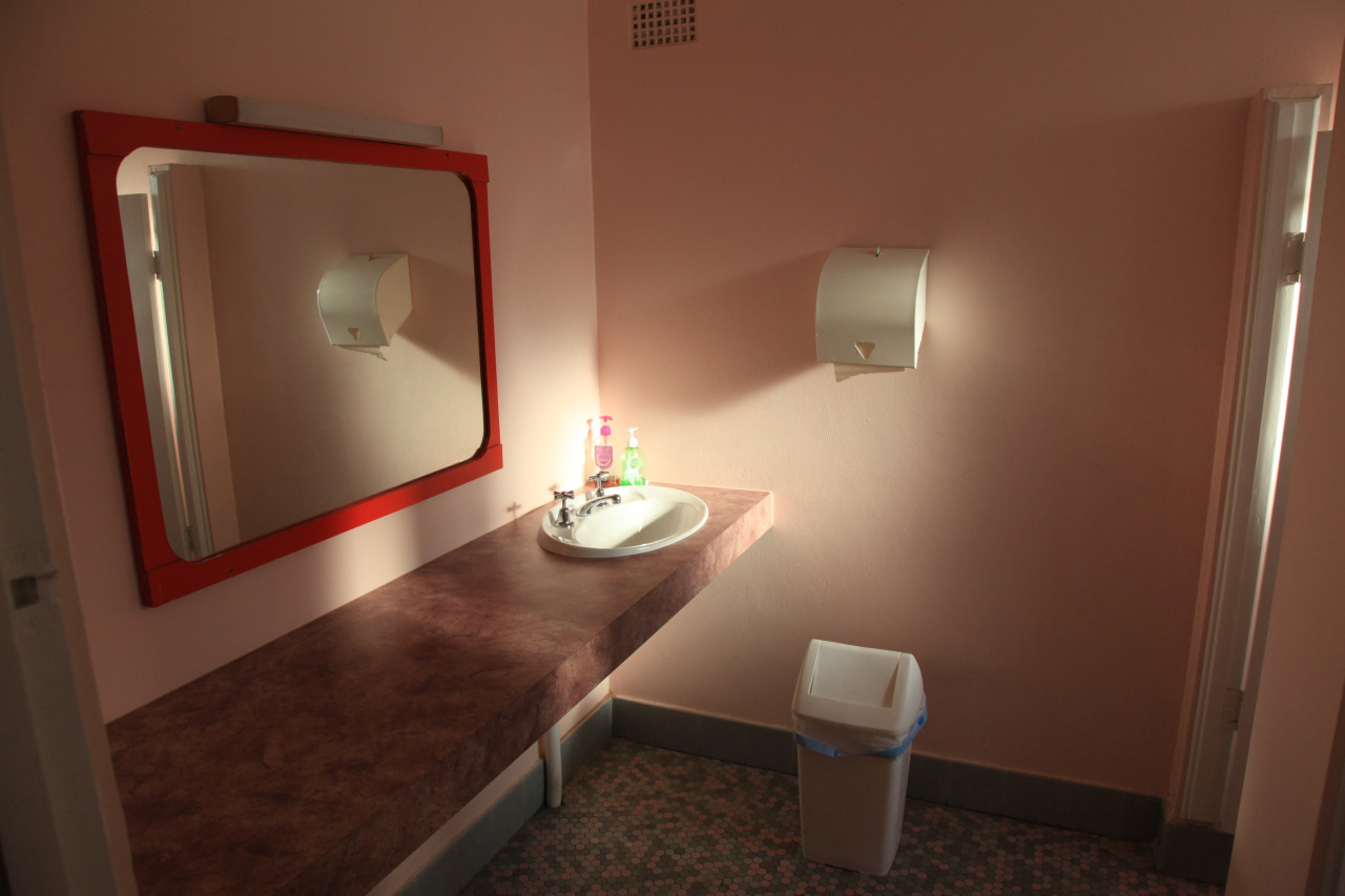 A well-lit bathroom features a pink wall, a red-framed mirror, a white sink with toiletries, a paper towel dispenser, and a trash can.