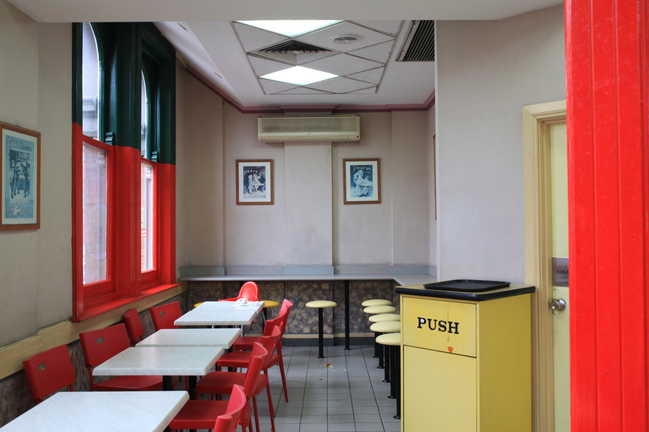 Interior view of an empty dining area with red chairs and white tables, framed posters on the neutral-colored walls, and overhead lighting.